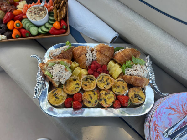 Silver tray of tuna-salad croissant sandwiches, mini quiches, strawberries, raspberries and pineapple slices beside a charcuterie board and napkins on a boat bench — casual boat brunch platter
