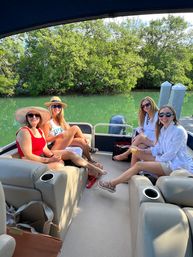 Four friends relaxing with drinks on a pontoon boat in a mangrove-lined inlet, wearing sun hats and sunglasses on a sunny summer day with green water, dock posts and outboard motor visible.