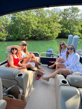 Four friends relaxing with drinks on a pontoon boat in a mangrove-lined inlet, wearing sun hats and sunglasses on a sunny summer day with green water, dock posts and outboard motor visible.
