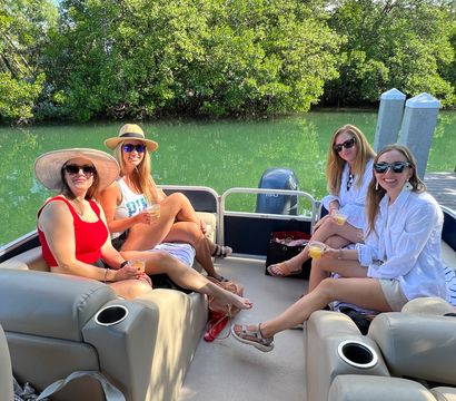 Four friends relaxing with drinks on a pontoon boat in a mangrove-lined inlet, wearing sun hats and sunglasses on a sunny summer day with green water, dock posts and outboard motor visible.
