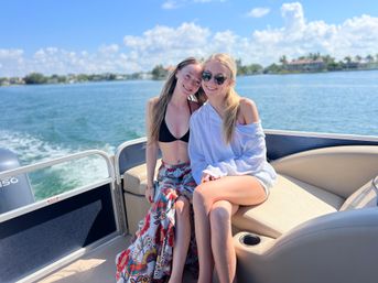 Two people smiling on a pontoon boat cruising a sunny coastal bay with waterfront homes, one in a black bikini top and floral skirt and the other in a white shirt and sunglasses.