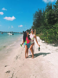 Two friends in bright swimsuits walking barefoot along a sunny tropical beach, holding drinks and smiling with turquoise water, anchored boats, and green shoreline foliage