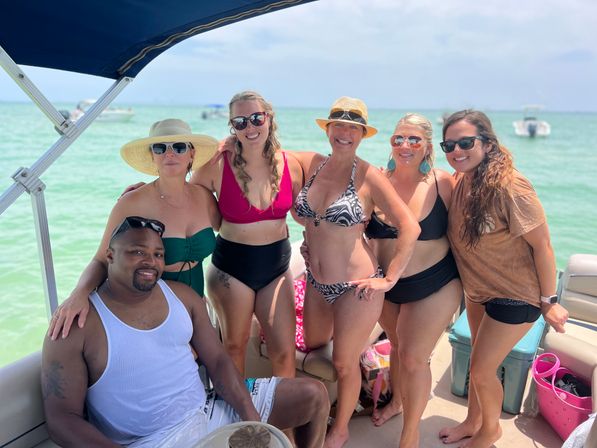 Six adults in swimsuits with sun hats and sunglasses posing and smiling on a pontoon boat over clear turquoise water on a sunny summer boat outing