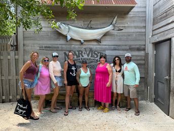 Beachside group photo of eight adults in summer clothes posing under a large mounted tarpon on a weathered wooden wall outside a coastal restaurant, sandy ground and leafy branches overhead.