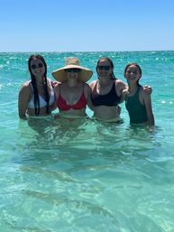 Four smiling women in swimsuits standing waist-deep in clear turquoise ocean water under a bright blue sky, one wearing a wide straw sunhat and sunglasses.