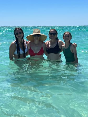 Four smiling women in swimsuits standing waist-deep in clear turquoise ocean water under a bright blue sky, one wearing a wide straw sunhat and sunglasses.