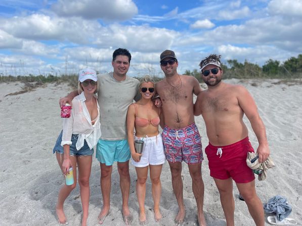 Five adults barefoot and smiling on a sandy beach under a blue sky with fluffy clouds, wearing swimsuits, shorts and sunglasses—casual summer beach scene.