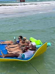Group of adults lounging on a yellow floating mat in turquoise ocean near a white-sand beach, wearing sunglasses and sun hats and holding drinks — sunny summer beach vibe