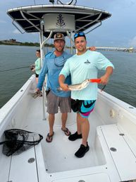 Two people on a center-console fishing boat holding a speckled trout they caught in coastal waters near a bridge, with fishing gear and a net on deck.