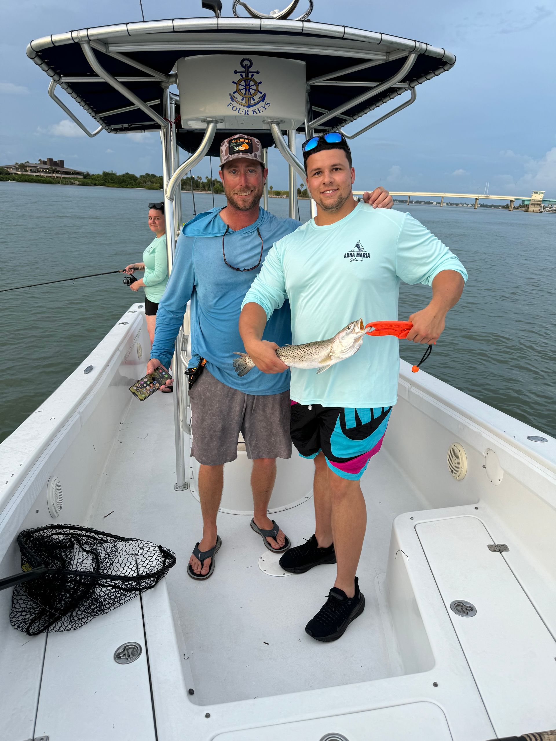 Two people on a center-console fishing boat holding a speckled trout they caught in coastal waters near a bridge, with fishing gear and a net on deck.