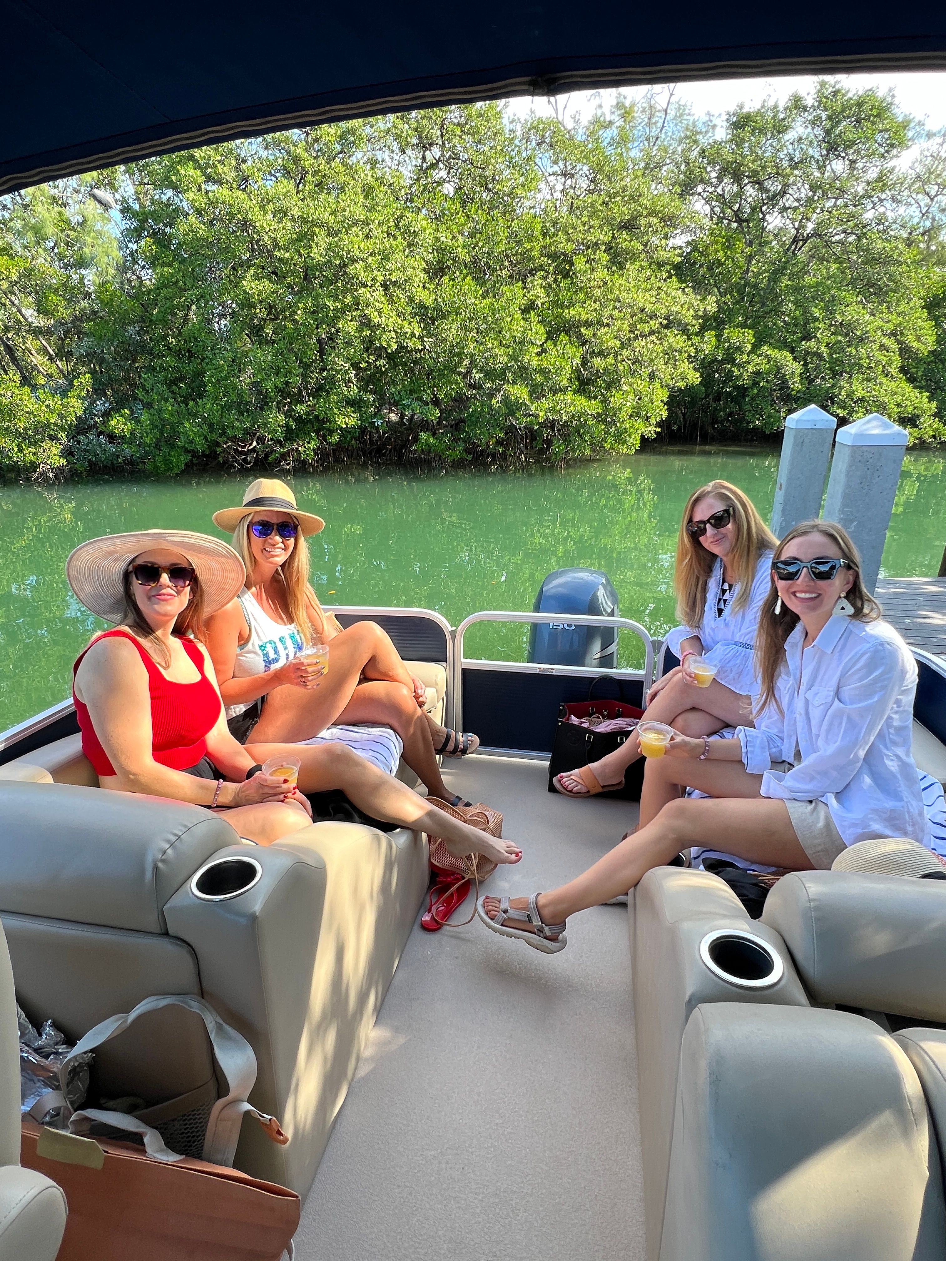 Four women in sun hats and sunglasses enjoying drinks and lounging on a pontoon boat docked along a mangrove-lined green inlet on a sunny day
