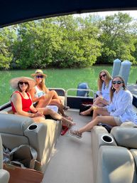 Four women in sun hats and sunglasses enjoying drinks and lounging on a pontoon boat docked along a mangrove-lined green inlet on a sunny day
