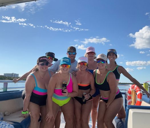 Eight adults in colorful swimsuits and slogan caps posing with canned drinks on a sunlit party boat, mirrored sunglasses, lifebuoy and calm coastal water under a bright blue sky.