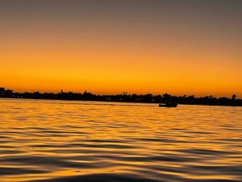Golden tropical sunset over a calm bay, silhouetted palm-lined shoreline and a small boat on rippling orange water.