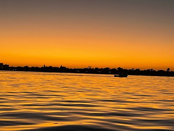Golden tropical sunset over a calm bay, silhouetted palm-lined shoreline and a small boat on rippling orange water.