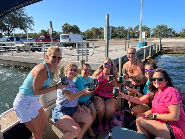 Eight women in colorful shirts and sunglasses toasting with drinks on a pontoon boat at a sunny marina dock with palm trees, parked trucks and calm water — lively coastal boat party scene.