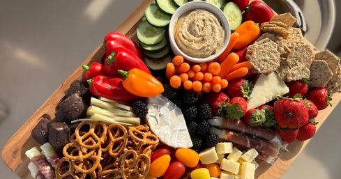 Colorful charcuterie grazing board on a wooden board with brie, cubed cheese, salami and prosciutto, pretzels, multigrain crackers, hummus, cucumber slices, mini sweet peppers, baby carrots, cherry tomatoes, strawberries, blackberries, and chocolate bites — fresh party platter.