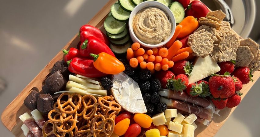 Colorful charcuterie grazing board on a wooden board with brie, cubed cheese, salami and prosciutto, pretzels, multigrain crackers, hummus, cucumber slices, mini sweet peppers, baby carrots, cherry tomatoes, strawberries, blackberries, and chocolate bites — fresh party platter.