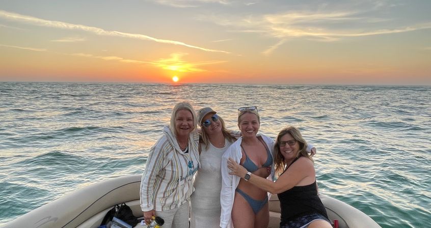 Four friends smiling together on a small boat during an ocean sunset, warm golden sun dipping to the horizon with rippling water and pastel sky