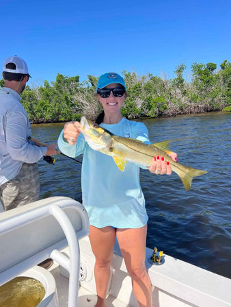 Smiling angler in a blue shirt and cap holding a freshly caught snook aboard a small boat with mangrove-lined inshore waters and clear blue sky.