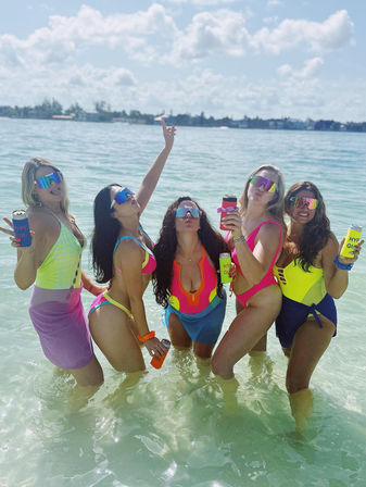 Five friends in neon swimsuits and mirrored visors posing waist-deep in clear turquoise water at a sunny tropical beach, holding colorful canned drinks