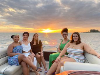 Five friends smiling on a pontoon boat at sunset, colorful clouds and golden sun reflecting across calm water.