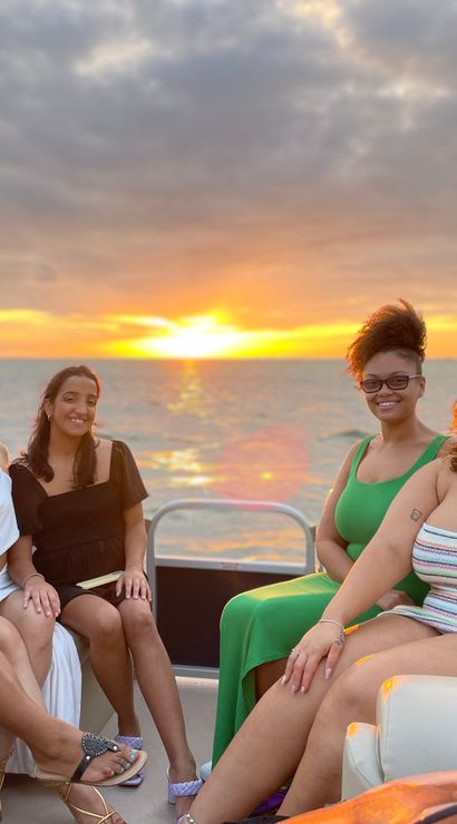 Five friends smiling on a pontoon boat at sunset, colorful clouds and golden sun reflecting across calm water.