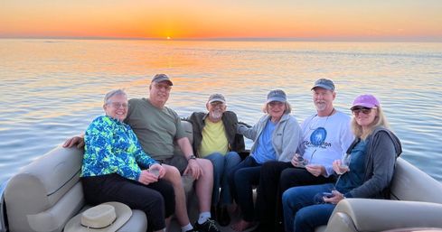 Six friends on a pontoon boat enjoying a vibrant sunset over a calm lake, smiling, wearing caps and holding wine glasses.