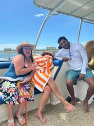 Three friends relaxing on a motorboat in calm coastal waters — woman in a straw hat and heart-shaped sunglasses giving a thumbs up, someone wrapped in an orange-and-white towel, and a smiling man in a white long-sleeve shirt and colorful swim trunks under a clear blue sky