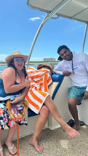 Three friends relaxing on a motorboat in calm coastal waters — woman in a straw hat and heart-shaped sunglasses giving a thumbs up, someone wrapped in an orange-and-white towel, and a smiling man in a white long-sleeve shirt and colorful swim trunks under a clear blue sky