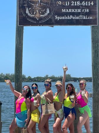 Six women in brightly colored neon swimsuits and reflective sunglasses posing on a wooden dock by a calm bay, holding tropical drinks and tote bags under a sunny blue sky.