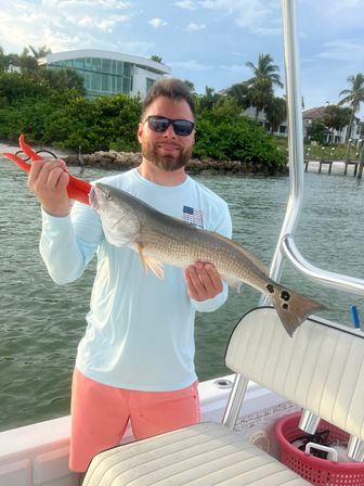 Angler on a boat holding a redfish (red drum) with black tail spots, inshore saltwater fishing near palm-lined coastal homes under a blue sky