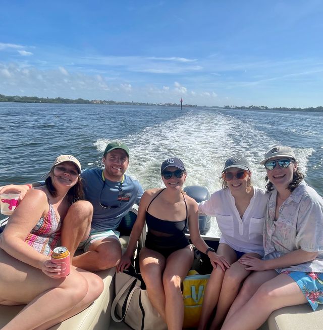 Five friends smiling on a motorboat, sitting by the engine wake under a bright blue sky with a coastal shoreline in the distance — sunny summer boating on open water.