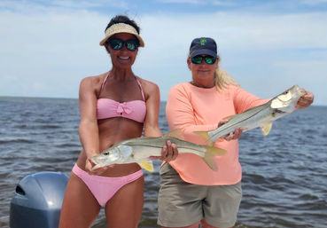 Two anglers on a boat proudly holding two snook they caught in calm inshore coastal waters under a sunny sky — inshore saltwater fishing catch