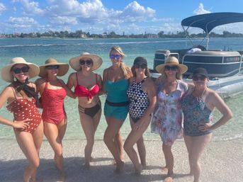 Seven women in colorful swimsuits and sun hats posing on a sunny beach with clear turquoise water and a pontoon boat offshore.