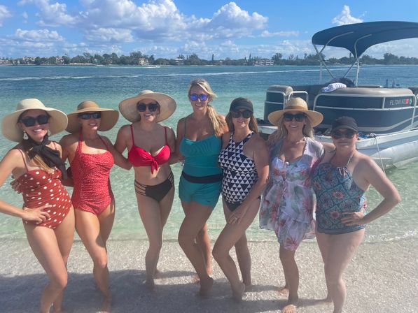 Seven women in colorful swimsuits and sun hats posing on a sunny beach with clear turquoise water and a pontoon boat offshore.