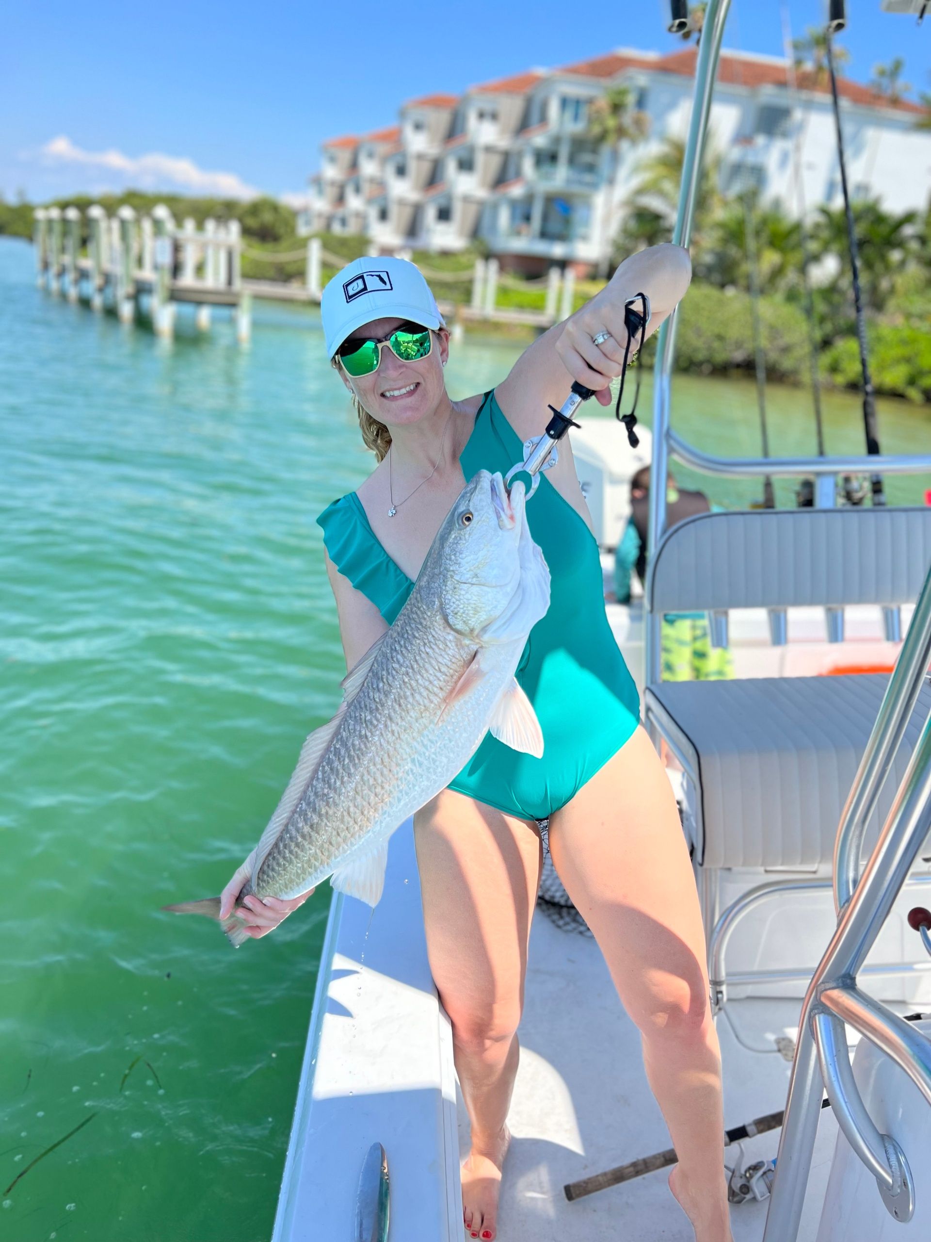 Smiling angler in a teal swimsuit and white cap holds a large silver fish with a lip gripper on a boat in turquoise coastal waters near a marina and waterfront condos.
