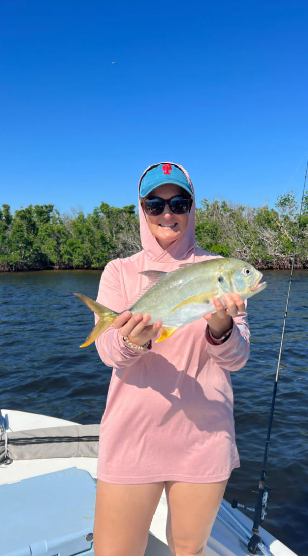 Smiling angler in a pink sun hoodie and blue cap holding a silver-yellow fish on a boat near mangrove-lined shore under a clear blue sky