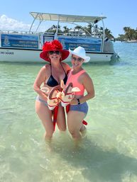 Two smiling women in colorful sun hats and swimsuits standing waist-deep in clear turquoise beach water, each holding an inflatable stick horse, with a charter boat and palm trees in the sunny background.