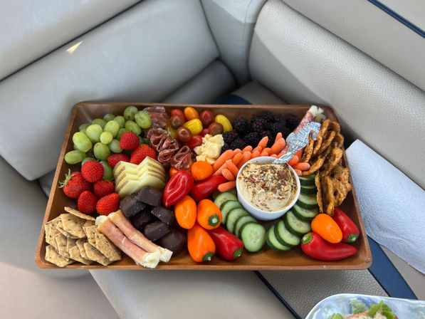 Colorful charcuterie grazing board on a wooden tray with grapes, strawberries, blackberries, sliced cucumber, baby carrots, mini sweet peppers, rolled cured meats, sliced cheese, dark chocolate bites, crackers, pretzels and a seasoned dip, set on a boat seat.