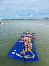 Smiling friends in swimsuits and straw hats lounging on a large blue floating mat in shallow coastal bay water, holding drinks with distant waterfront condos and boats under a sunny, partly cloudy sky