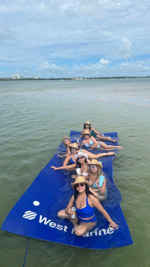 Smiling friends in swimsuits and straw hats lounging on a large blue floating mat in shallow coastal bay water, holding drinks with distant waterfront condos and boats under a sunny, partly cloudy sky