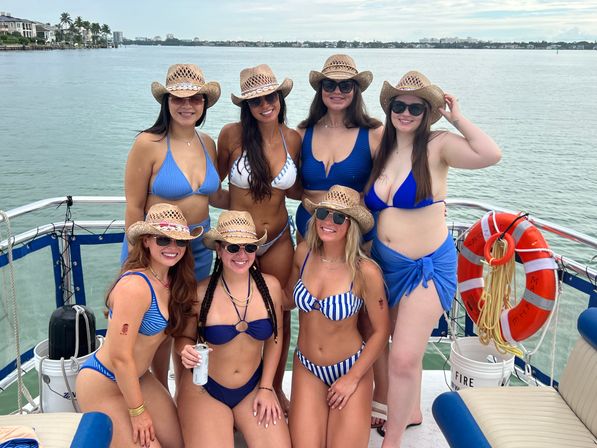 Seven friends in blue bikinis and straw cowboy hats smiling on a boat deck with sunglasses, life preserver, and coastal skyline over calm bay water