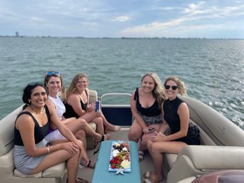 Five smiling friends relaxing on a pontoon boat over open water, holding drinks with a colorful charcuterie board on the center table under a cloudy daytime sky.