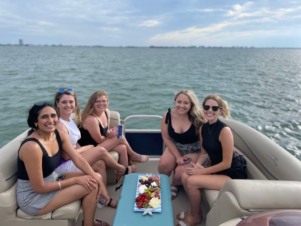 Five smiling friends relaxing on a pontoon boat over open water, holding drinks with a colorful charcuterie board on the center table under a cloudy daytime sky.