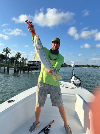 Angler on a small boat holding a large snook with a lip gripper, dock and palm trees along a sunny Florida coast under blue sky and scattered clouds.
