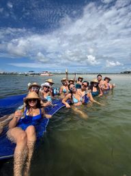 Group of friends lounging on a blue floating mat at a shallow sandbar, wearing straw hats and sunglasses and holding drinks, with boats and a coastal skyline in the background under a bright partly‑cloudy sky.