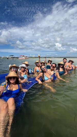Group of friends lounging on a blue floating mat at a shallow sandbar, wearing straw hats and sunglasses and holding drinks, with boats and a coastal skyline in the background under a bright partly‑cloudy sky.