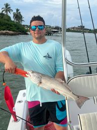Angler on a boat holding a freshly caught redfish with a lip-grip, wearing sunglasses and a light blue Anna Maria Island shirt, with palm trees, docks and coastal waters in the background