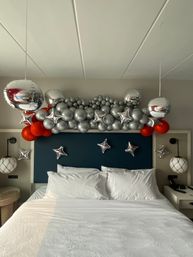Hotel room bed with white linens and teal headboard decorated with a silver-and-red balloon garland and metallic star-shaped and reflective orb balloons overhead